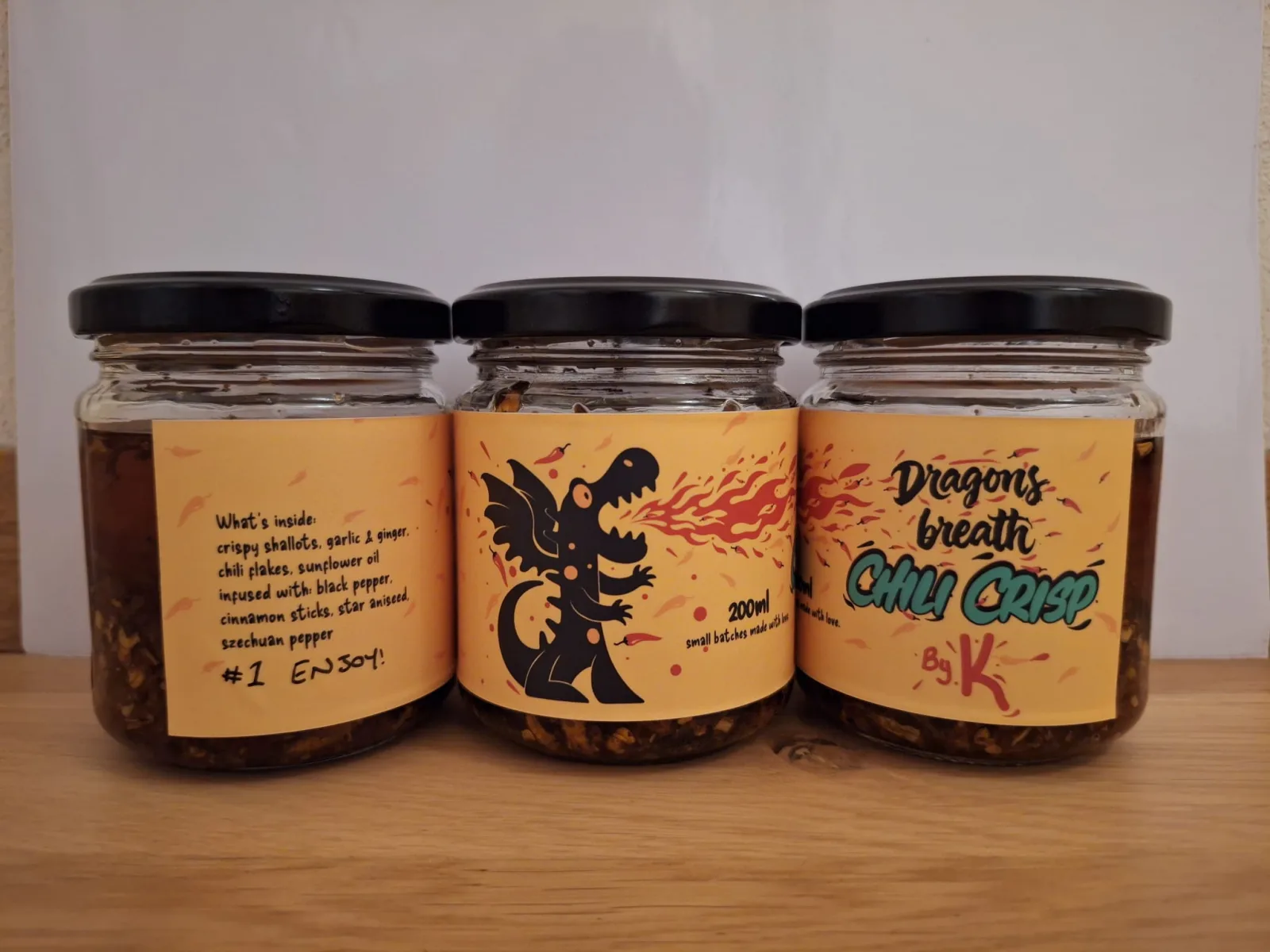 Three jars of Dragon's Breath Chili Crisp by K on a wooden table, labels showing a black dragon breathing fire